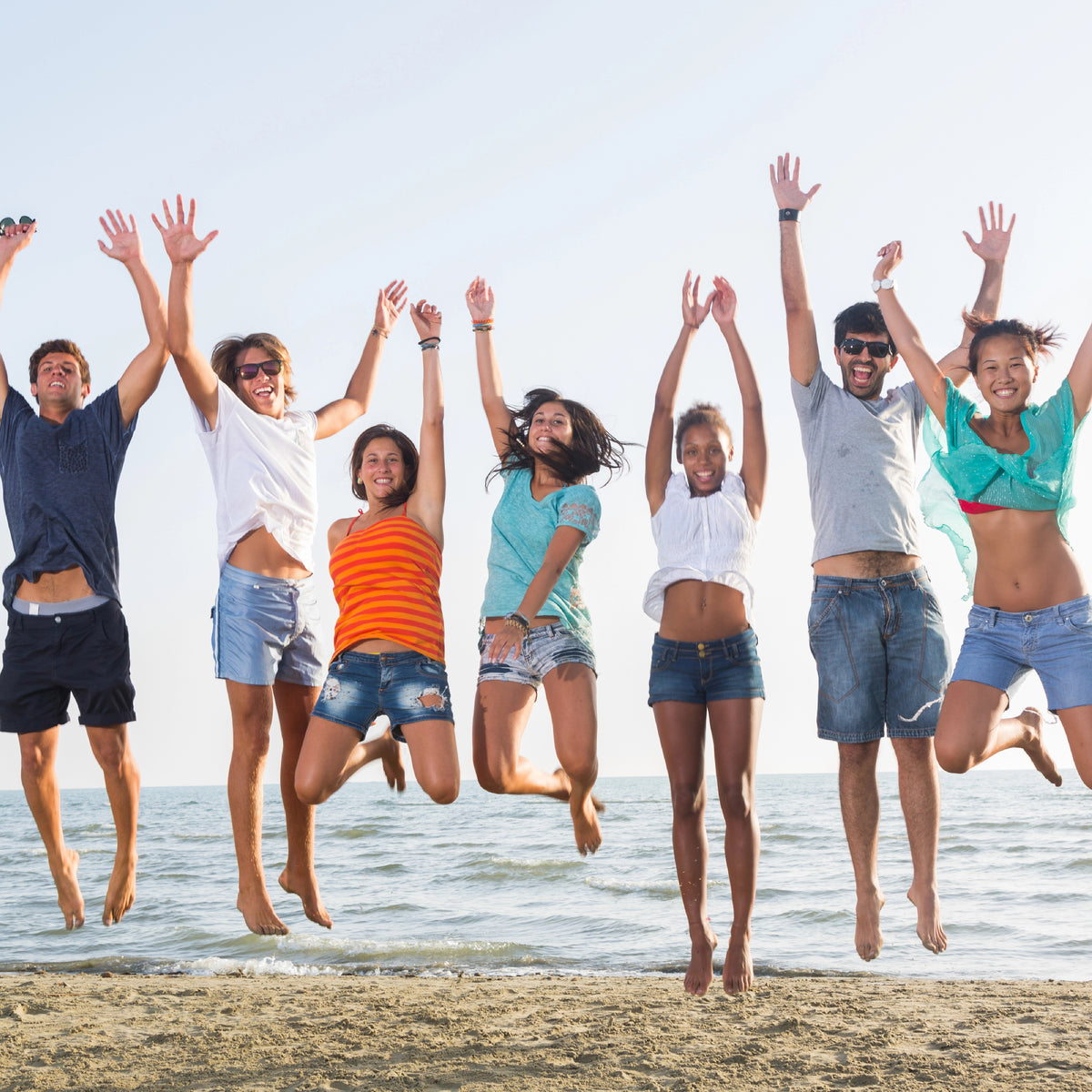 A group of seven young adults jumping happily on a sandy beach with the ocean and clear sky in the background.